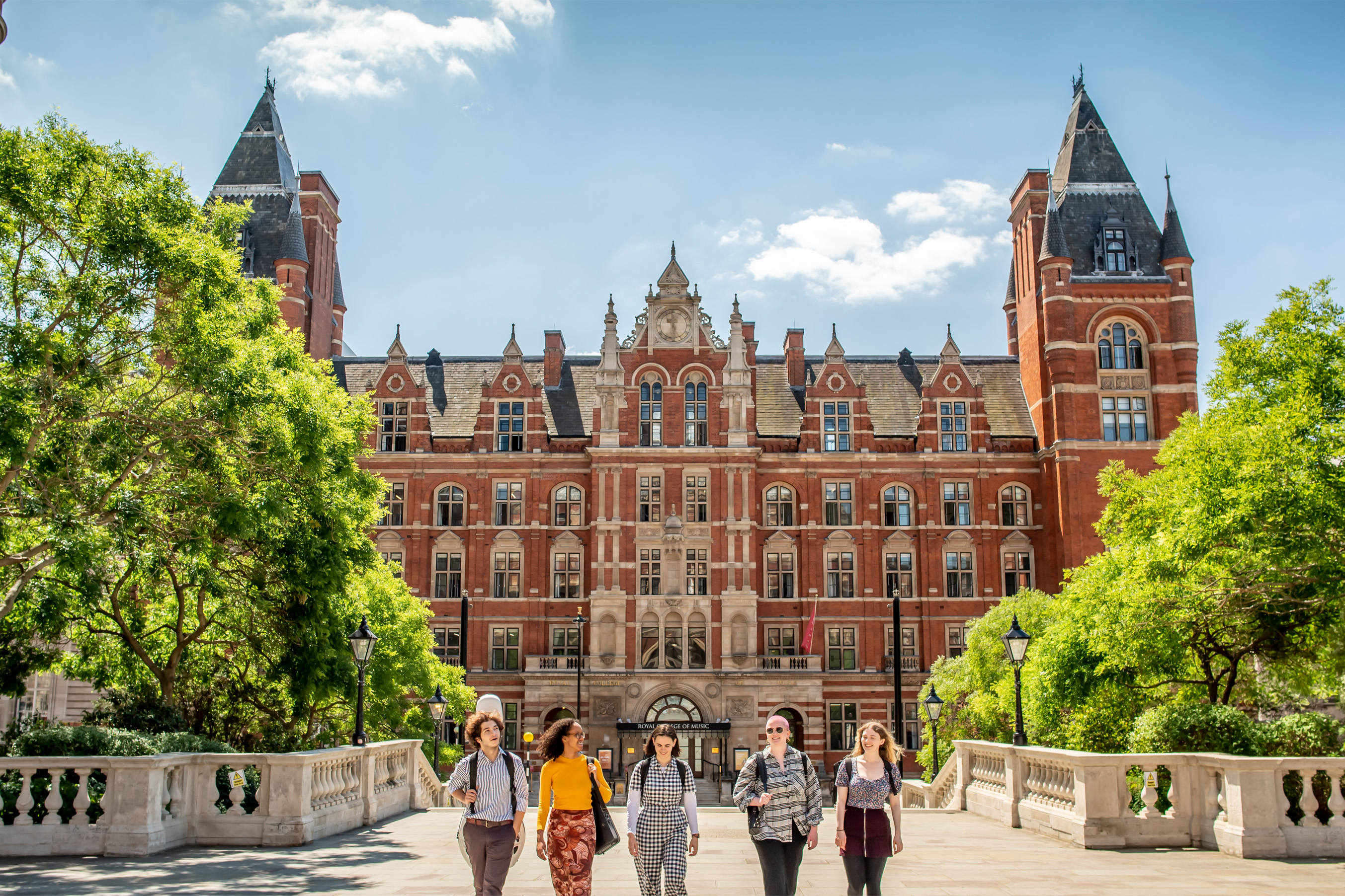 A group of students wearing casual clothes, walking towards the front entrance of the Royal College of Music, an old, red brick, Victorian building, in the background.